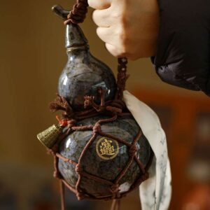 A person holding an artisanal ceramic gourd water bottle with a dark, kiln-fired glaze, secured in a hand-tied brown rope carrier with a small brass bell.