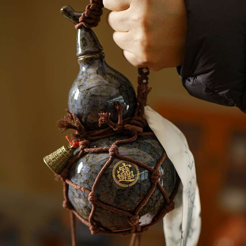 A person holding an artisanal ceramic gourd water bottle with a dark, kiln-fired glaze, secured in a hand-tied brown rope carrier with a small brass bell.